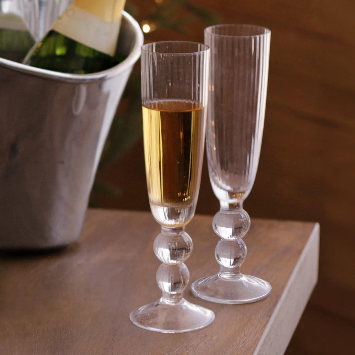 Two elegant Beatriz Ball "Venice" champagne glasses, one filled with sparkling wine and one empty, rest on a wooden table beside a metal ice bucket holding a bottle—an exquisite set of four from Beatriz Ball, perfect for special celebrations.