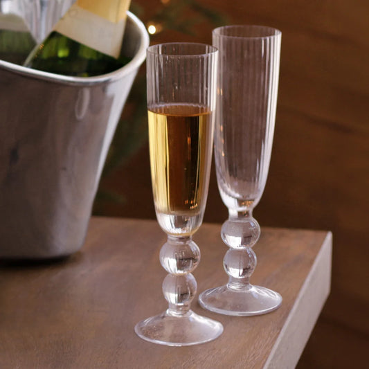 Two elegant Beatriz Ball "Venice" champagne glasses, one filled with sparkling wine and one empty, rest on a wooden table beside a metal ice bucket holding a bottle—an exquisite set of four from Beatriz Ball, perfect for special celebrations.