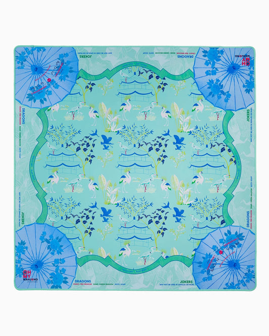 The Oh My Mahjong Veranda Mat in Tube by Oh My Mahjong is an elegant mahjong table mat showcasing a light blue and green floral pattern, highlighted by graceful cranes, pagodas, and four blue decorative umbrellas in the corners. With clearly labeled dragons and jokers in each corner, this mat combines beauty and function to elevate your next game night.