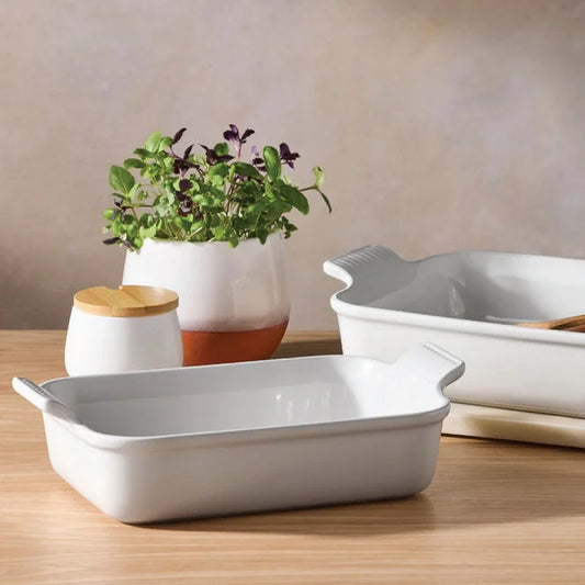 Two Le Creuset 2.5 qt Heritage Rectangular Baking Dishes in white rest on a wooden table, with a small potted plant and a white jar with a wooden lid in the background.